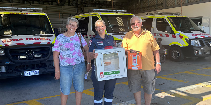 A paramedic holding an AED stands outside an ambulance branch with two committee members